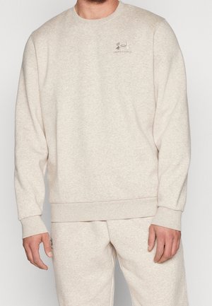 Mann trägt ein hellbeiges Under Armour Sweatshirt und passende Jogginghose, steht mit entspannt an den Seiten hängenden Armen.