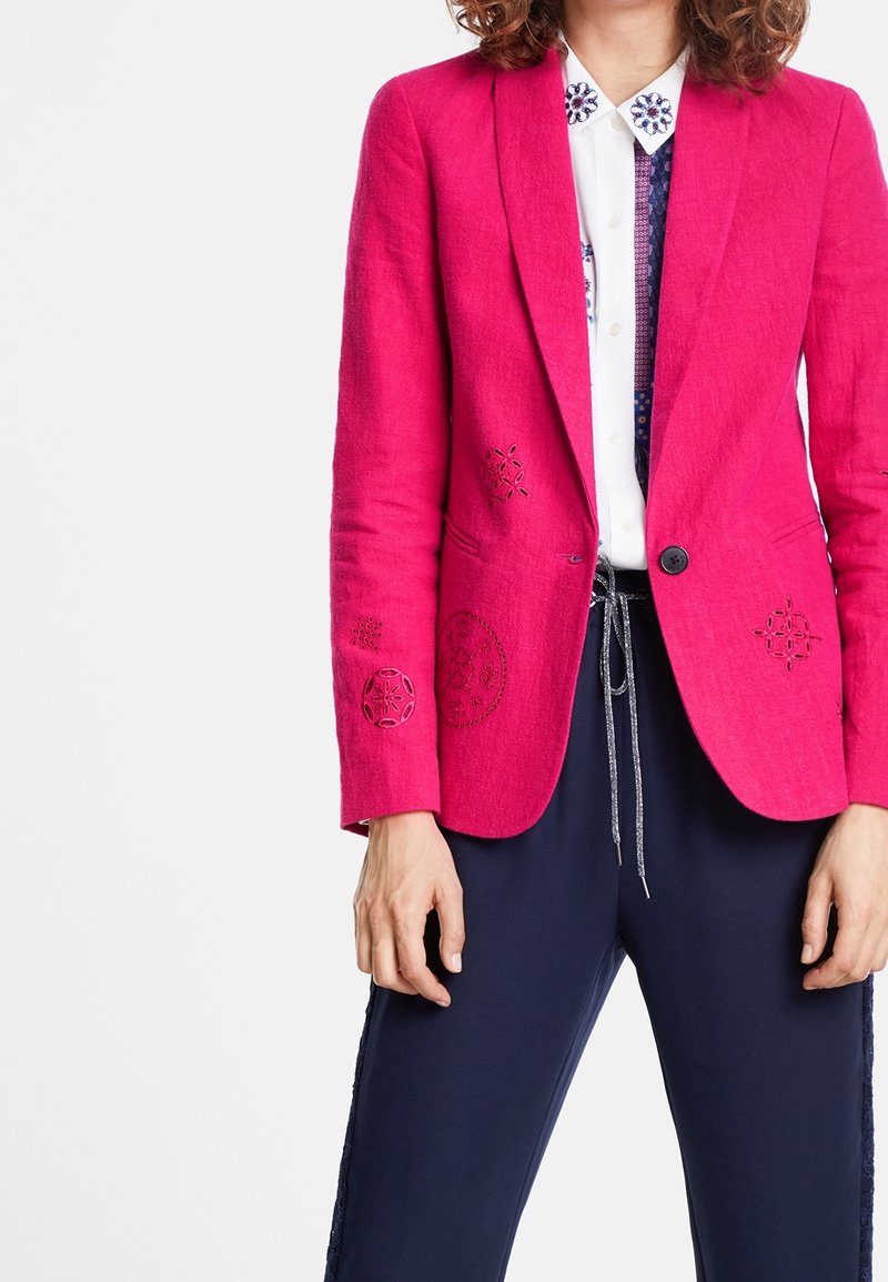 Femme portant un blazer rose vif avec des motifs brodés sur une chemise blanche et un pantalon bleu foncé avec taille à cordon.