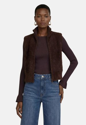 Kvinde iført en mørkebrun fluffy vest over en brun ribbet langærmet top og blå jeans, stående med den ene hånd på hoften.