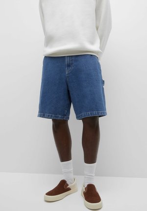 Man draagt blauwe denimshorts, wit overhemd met lange mouwen, witte sokken en bruine slip-on sneakers, staand tegen een effen witte achtergrond.