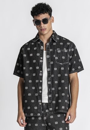 Muž nosiaci čiernu krátku košeľu na gombíky a zodpovedajúce šortky s bielym vzorom loga, biele tričko a čierne slnečné okuliare.