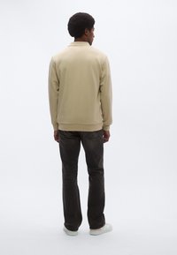 Bézs pulóver magas nyakkal, bordázott mandzsettákkal és szegéllyel, sötét farmerrel és világos sportcipővel párosítva. Egyszerű, minimalista dizájn.