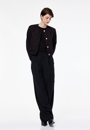 Donna in piedi con le mani dietro la schiena, che indossa un cardigan nero testurizzato con bottoni, pantaloni neri larghi e scarpe nere.