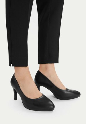 Persona vistiendo pantalones negros ajustados y zapatos de piel negros de tacón medio, de pie sobre un fondo claro.