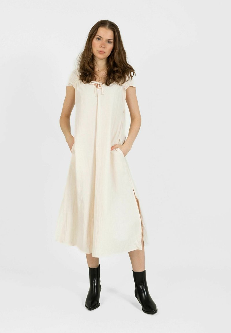 Jeune femme en robe midi crème à manches courtes et fentes latérales, portant des bottines noires, debout avec les mains dans les poches.