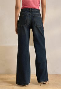 Mörka denimbyxor med vida ben och hög midja, som har två bakfickor och en ren fåll. Modellen bär ljusa klackar.