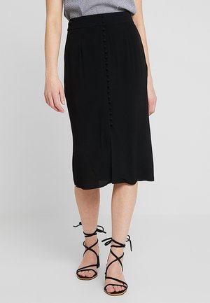 Vrouw draagt een zwarte midi-rok met bedekte knopen aan de voorkant, gecombineerd met zwarte sandalen met hak en bandjes, en een gestreept topje.