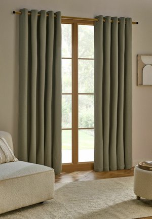 Grüne Vorhänge hängen an einer Holzstange über einer hohen Glastür mit einem hellbeigen Sofa und einem texturierten Teppich in einem neutralfarbenen Wohnzimmer.