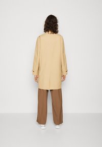 Beige op maat gemaakt jasje met lange mouwen en een achteropening, gecombineerd met brede bruine broeken en witte sneakers. Glad textuur van de stof.