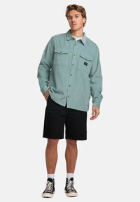Lys tealfarvet kordfløjspænde med to frontlommer, knaplukning og et sort logo-patch, parret med sorte shorts og sorte høje sneakers.