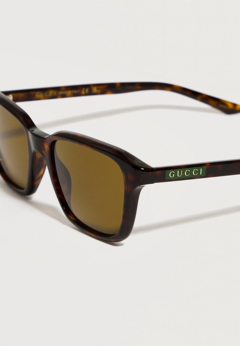 Square tortoiseshell sunglasses with brown lenses and a Gucci logo plaque on the temples resting on a white surface.