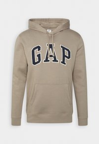 Beżowa bluza z kapturem z napisem "GAP" w granatowych literach, kapturem ze sznurkiem, kieszenią kangurka oraz prążkowanymi mankietami i dolnym brzegiem. Wykonana z miękkiej bawełnianej mieszanki.