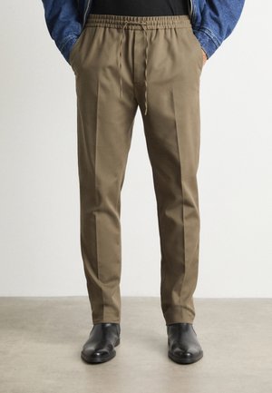 Man draagt olijfgroene, op maat gemaakte broek met trekkoord en voorplooien, gecombineerd met zwarte leren laarzen op een grijze vloer.