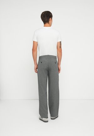 Mann mit dem Rücken zur Kamera, trägt ein schlichtes weißes Kurzarm-T-Shirt, graue gerade geschnittene Hosen und graue und weiße Sneakers, vor weißem Hintergrund.