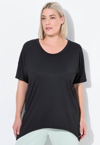 Černý top s krátkým rukávem a širokým výstřihem ve tvaru U, volný střih, měkká textura a zakulacený lem. Modelka má na sobě světle zelené kalhoty.