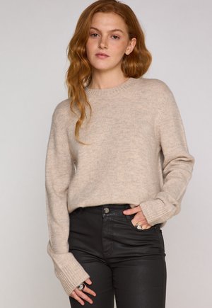 Beiger Strickpullover mit rundem Ausschnitt und gerippten Bündchen, kombiniert mit schwarzen, figurbetonten Hosen. Die Textur wirkt weich und leicht flauschig.