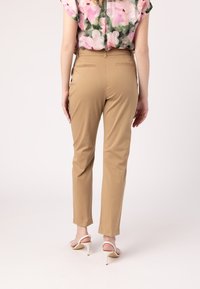 Béžové nohavice na mieru so slim fit strihom, hladkou textúrou, jemnými zadnými vreckami, skombinované s bielymi sandálmi na vysokom podpätku. Kvetinový top nie je zobrazený.
