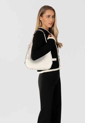 Sac à bandoulière en cuir blanc avec motif en losanges, quincaillerie dorée et bandoulière chaîne, tenu par une personne portant un pull noir et un pantalon.
