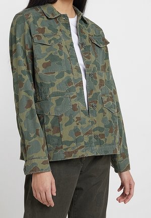 Person iført en grøn og brun camouflage knapjakke over en hvid skjorte, kombineret med mørke bukser, stående mod en neutral baggrund.