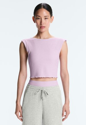 Jeune femme aux cheveux foncés portant un haut court sans manches lilas et un pantalon de jogging gris clair avec cordon sur un fond clair uni.