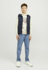 Mornarsko puffer brezrokavnik s horizontalnimi šivi, nošen čez svetlo sivo kapuco z grafiko, v kombinaciji s svetlo modrimi jeans hlačami in belimi teniskami.
