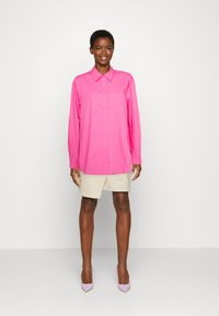 Camisa de mangas longas com botão, em rosa vibrante, feita de um tecido suave, com colarinho e um único bolso no peito. Combinada com calções bege.