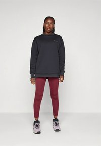 Sudadera negra acolchada con cuello redondo y puños acanalados, combinada con leggings burdeos. El modelo lleva zapatillas deportivas grises y negras.