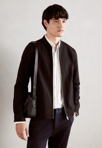 Μαύρο φερμουάρ μπουφάν πάνω από άσπρο πουκάμισο με κουμπιά, συνδυασμένο με σκούρους παντελόνες. Το μοντέλο κρατά μια μικρή μαύρη τσάντα χιαστί.