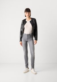 Svart skinnjakke med rund krage, tettsittende grå jeans og hvite sneakers. Modellen står mot en vanlig hvit bakgrunn.