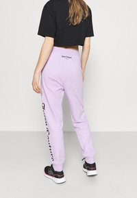 Lilacové tepláky s elastickými manžetami, se znakem na pasu a po straně, doplněné černým zkráceným tričkem a sportovními botami.