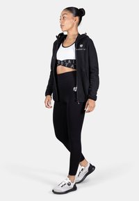 Black zip-up jacket over a white sports bra, paired with high-waisted black leggings. White and black sport shoes with a unique fastening system.