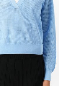 Ljusblå stickad tröja med V-ringning, som har en nätstruktur och ribbade ärmslut, i kombination med en svart veckad kjol.