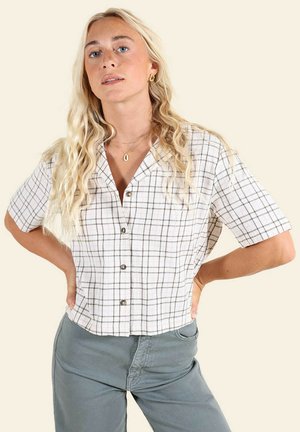 Femme blonde aux cheveux ondulés portant une chemise à carreaux blanche à manches courtes et un pantalon gris, debout les mains sur les hanches devant un fond crème.