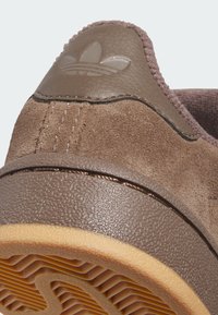 Brown suede sneaker with a textured brown rubber sole. Features a smooth brown leather accent with embossed logo and contrasting stitching.