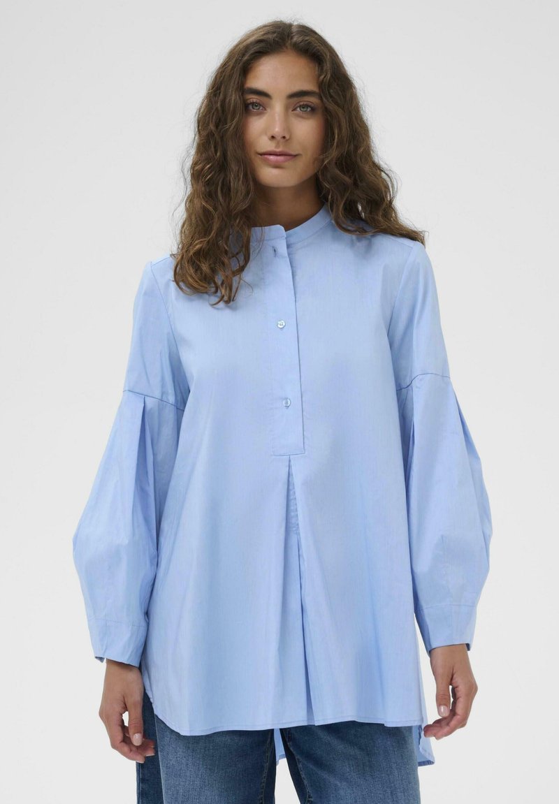 Frau mit langem, lockigem braunem Haar, die eine lockere hellblaue Knopfleiste-Bluse mit voluminösen Ärmeln und eine blaue Jeans trägt, vor einem einfarbigen Hintergrund.