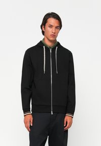 Uomo che indossa una felpa nera con zip e cappuccio grigio, e pantaloni neri, in piedi di fronte a uno sfondo bianco semplice.