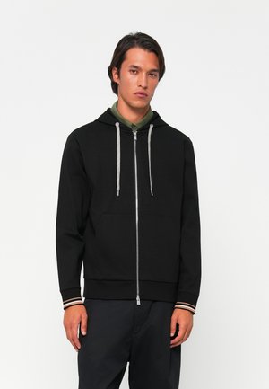 Man wearing black zip-up hoodie with grey drawstrings and black pants, standing against a plain white background.