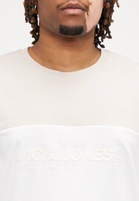 Beige en witte T-shirt met korte mouwen, met reliëf verworpen "JACK & JONES" tekst op de borst, zachte stof en contrasterende kleurvlakken.