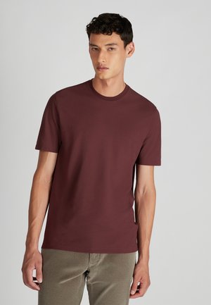 Jonge man in een effen bordeauxrode t-shirt met korte mouwen en lichtbruine ribfluwelen broek, staande tegen een effen lichte achtergrond.