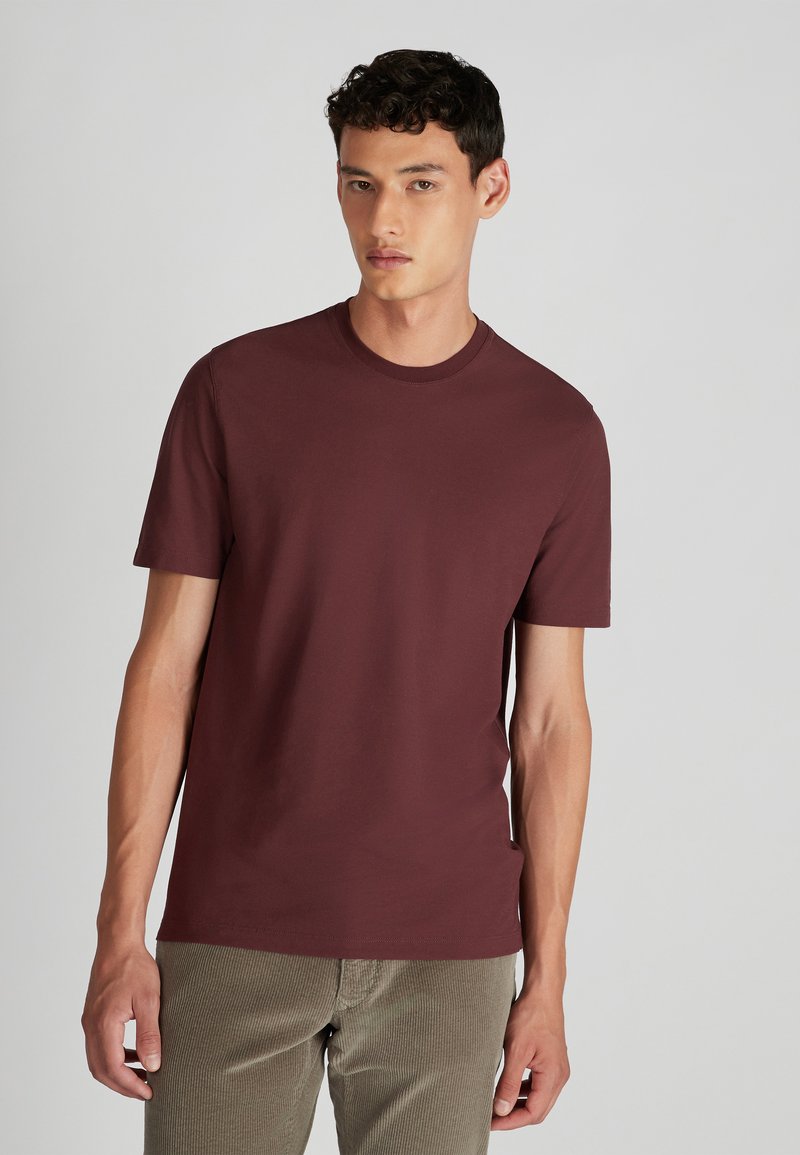 Un jeune homme portant un t-shirt court à manches courtes de couleur bordeaux uni et un pantalon en velours côtelé beige clair, se tenant devant un fond clair.