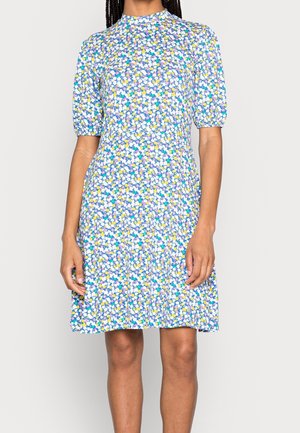 Mujer con un vestido de manga corta, largo hasta la rodilla, con un escote alto y un pequeño patrón floral colorido sobre un fondo azul.