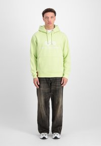 Felpa verde chiaro con tasca frontale e cordini, con un logo bianco. Indossata con jeans scuri e strappati e scarpe da ginnastica bianche.