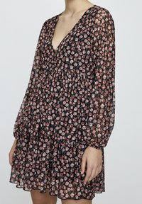 Robe noire à fleurs avec manches longues en voile transparent, décolleté en V profond, jupe évasée à volants superposés, et petits motifs floraux roses sur tout le tissu.