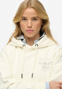 Hoodie crème avec cordons et poche avant, arborant le logo "Superdry & Co". Col rayé visible en dessous. Texture douce et lisse.