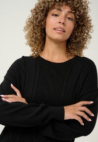 Fekete kötött pulóver, textúrázott mintákkal, bordás nyakkal és mandzsettákkal, valamint széles ujjal. Az anyag puha, kényelmes viseletet biztosít.