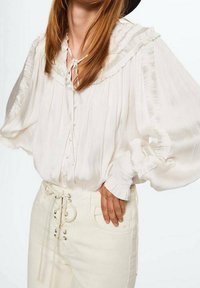 Blouse blanche légère avec col à volants, texture plissée et fermeture à boutons. Associée à un pantalon blanc taille haute avec détail à lacets.