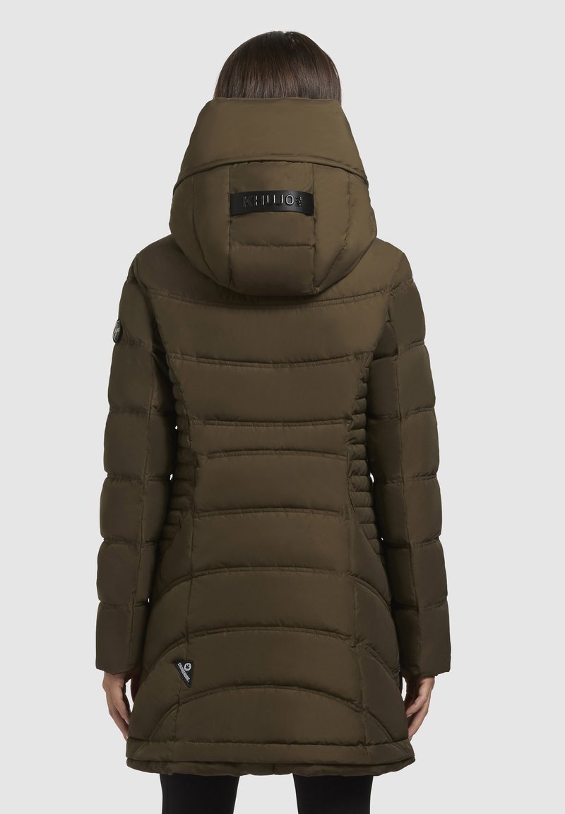 Frau in einem langen, olivgrünen, gesteppten Puffer-Coat mit großer Kapuze, die mit dem Rücken zu einer schlichten grauen Hintergrund steht.