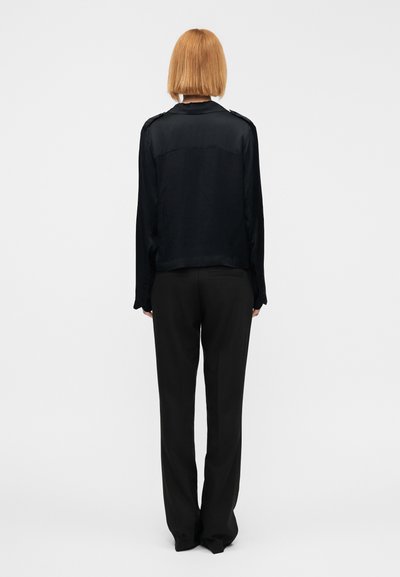 Μαύρο πουκάμισο με μακριά μανίκια, ομαλή υφή και επιθετικά στα ώμους, σε συνδυασμό με μαύρο, άνετο παντελόνι. Ουδέτερο υπόβαθρο.