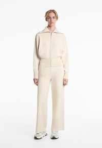 Ensemble de survêtement en tricot couleur crème avec un haut zippé à large col, et un pantalon droit ample. Porté avec des baskets grises et blanches.