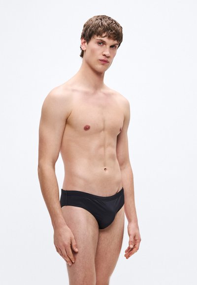 Cuecas de banho pretas com textura lisa, design minimalista e cintura elástica, usadas por um modelo com expressão neutra contra um fundo simples.